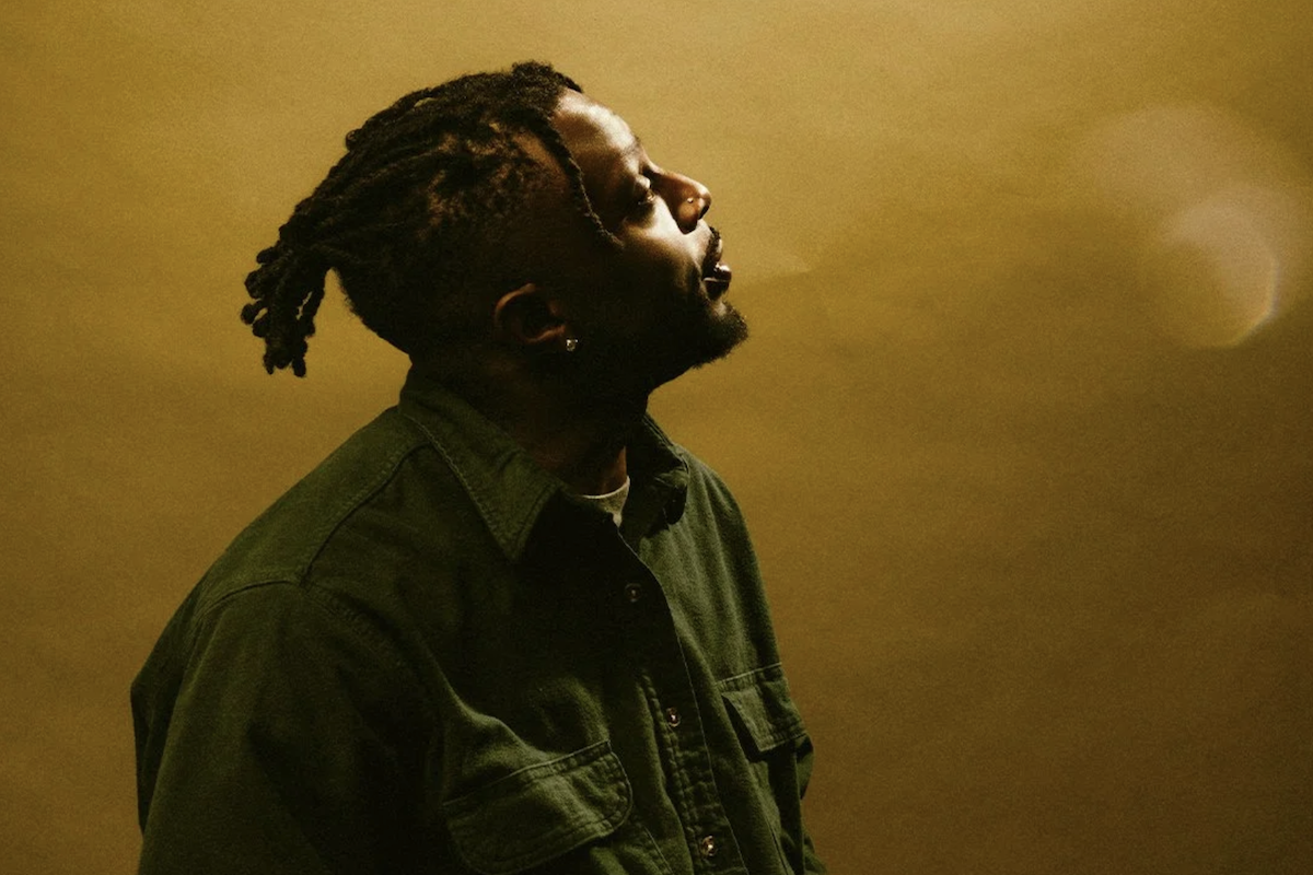Profile portrait of rapper Isaiah Rashad in a green jacket looking upward against a warm yellow backdrop.